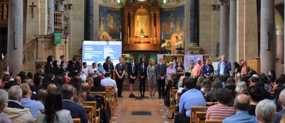 A line of school students, parents and teachers stand at the back of a church, in front of a large crowd of people sitting and watching from the pews, during a Refugee Welcome Schools' celebration in Leicestershire.