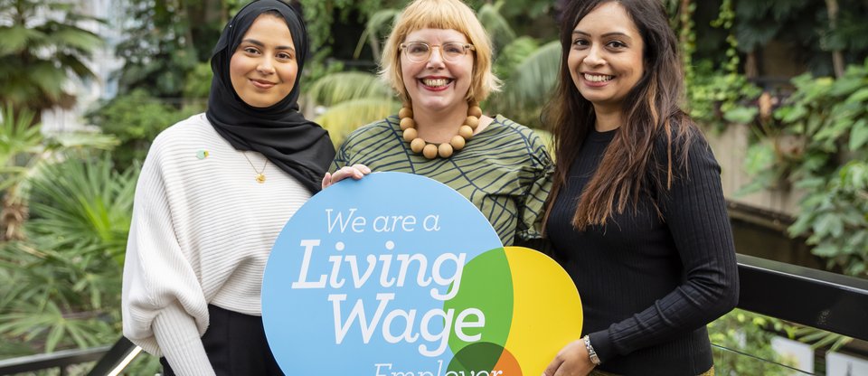 Three women stand smiling facing the camera, holding a Living Wage Foundation employer plaque. They are surrounded by green plants and the environment is fresh and bright.