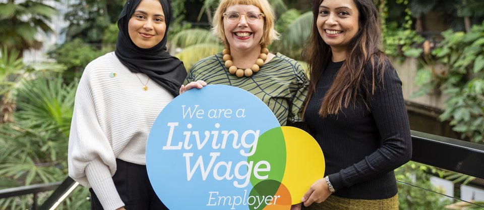 Three women stand smiling facing the camera, holding a Living Wage Foundation employer plaque. They are surrounded by green plants and the environment is fresh and bright.