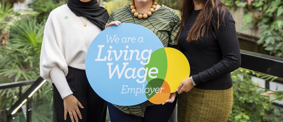 Three women stand smiling facing the camera, holding a Living Wage Foundation employer plaque. They are surrounded by green plants and the environment is fresh and bright.