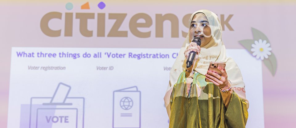 Woman stands with microphone in front of projector with information about the Citizens UK Voter Registration Championsscheme