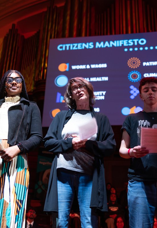 A Citizens UK campaigner is sharing her testimony on stage with a group of five people.