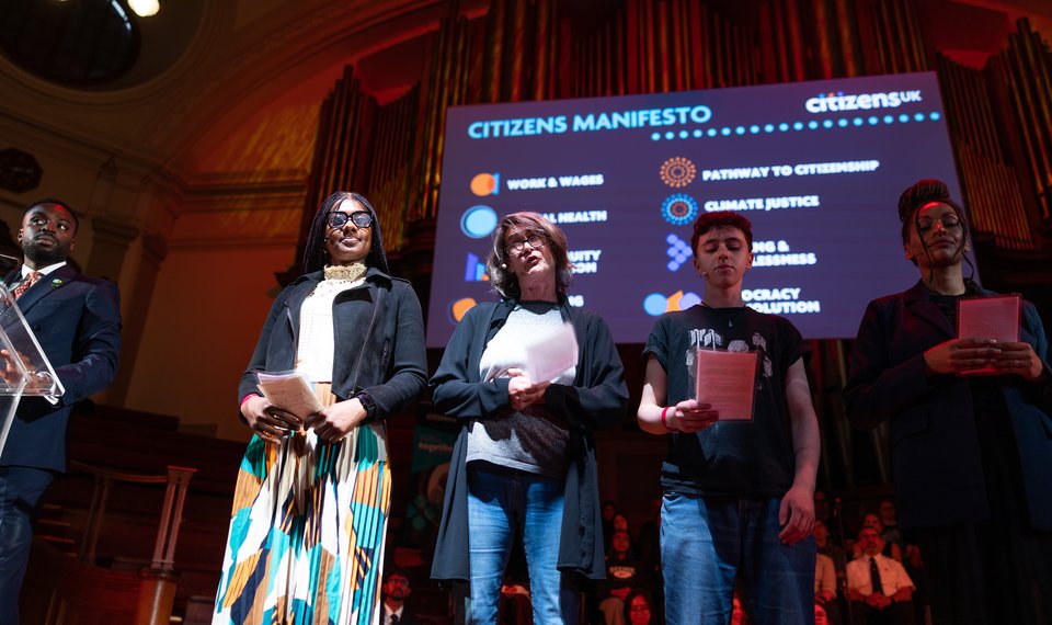 A Citizens UK campaigner is sharing her testimony on stage with a group of five people.