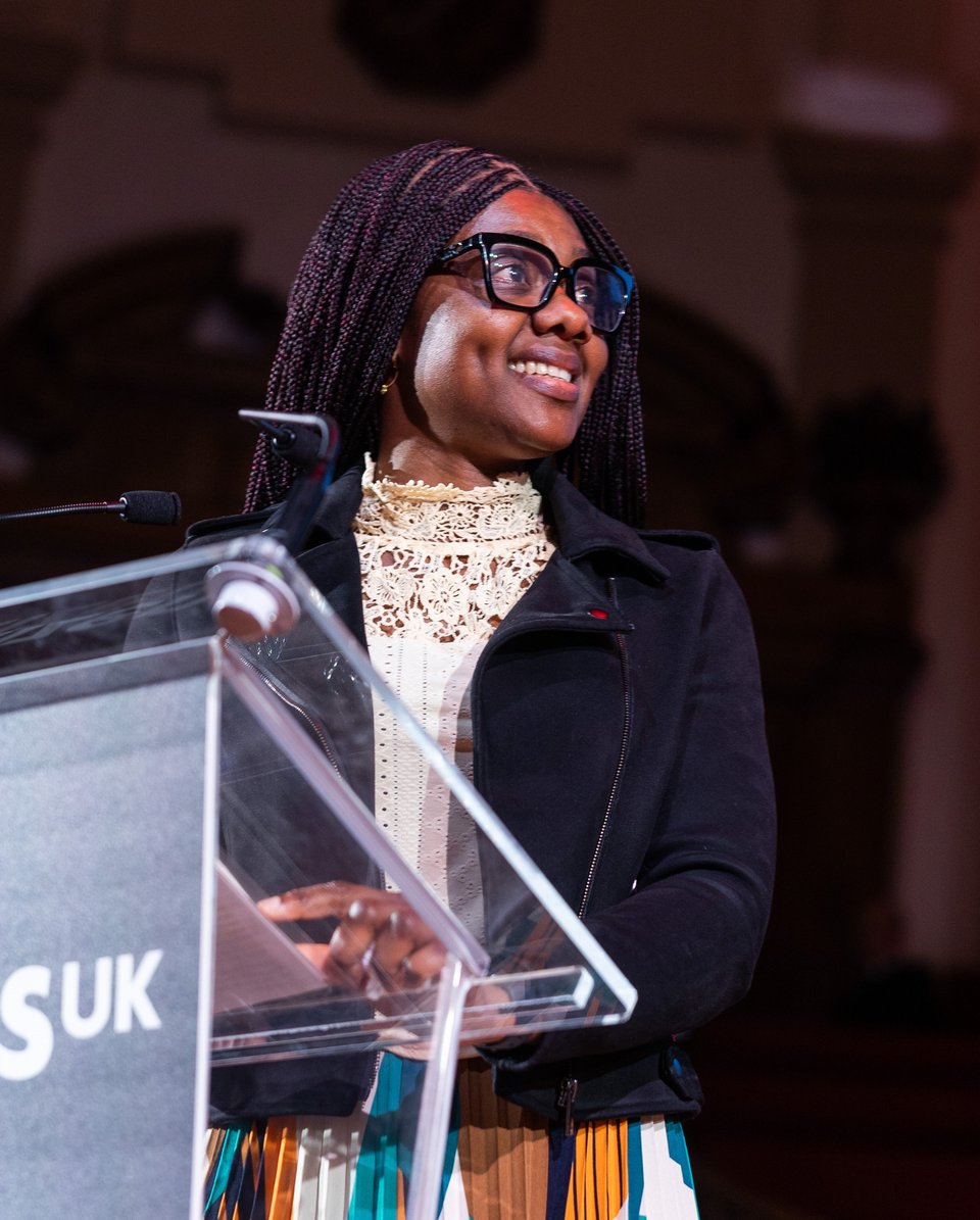 A Citizens UK leader is smiling and speaking on stage at a Citizens UK assembly