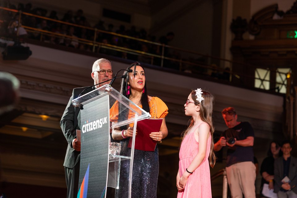 A Citizens UK leader asking a question on stage to Angela Rayner with her young daughter.