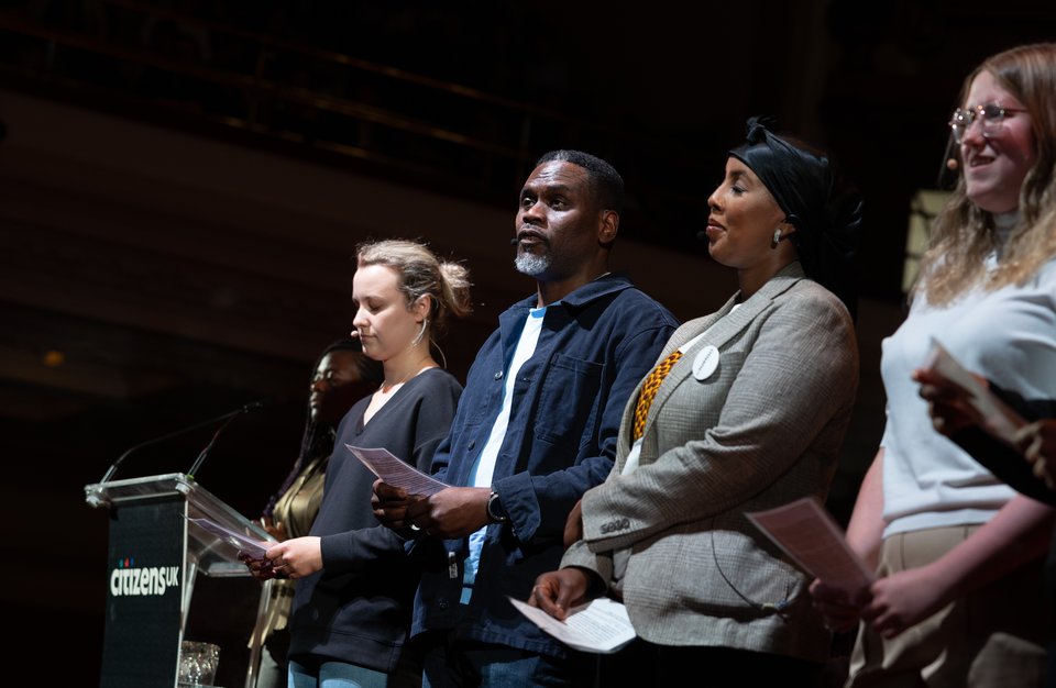 A Citizens UK leader on stage, sharing testimony amongst a group of people