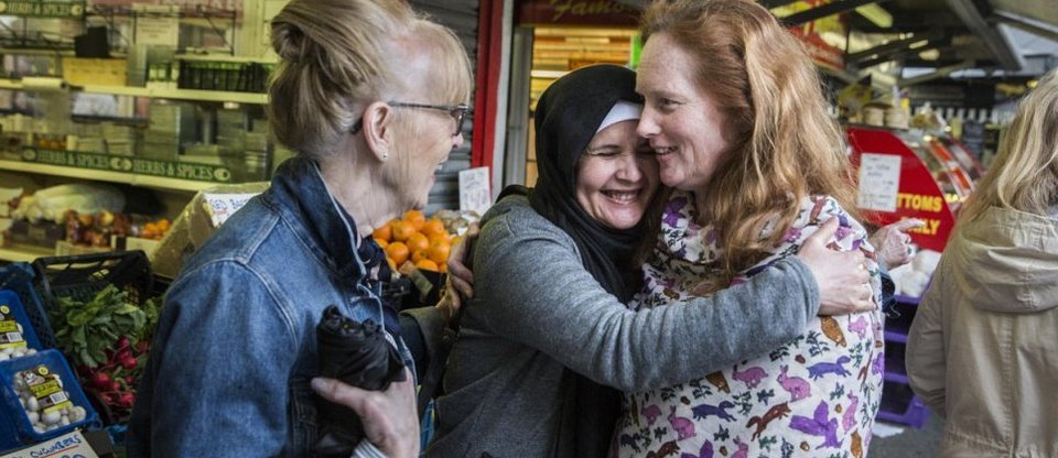 Three women involved in a Sponsor Refugees welcome group are outside a supermarket after some grocery shopping. The woman on the left is smiling and looking to her left, whilst the woman in the middle and on the right are laughing and hugging.