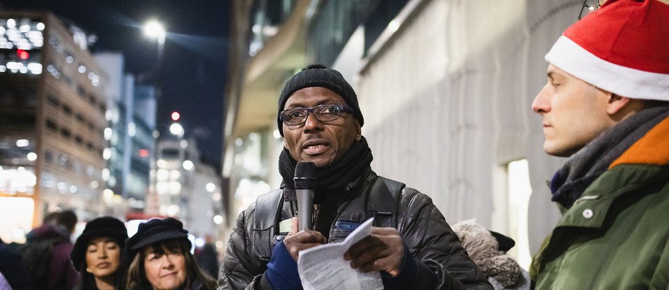 Community leader speaking outside a business, calling for the real Living Wage. It is night time in winter. People are wearing hats and gloves and are illuminated by street lights.