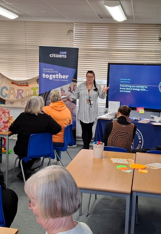 A woman standing in front of a TV screen and speaking to a room of people around tables.