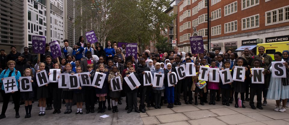 A large group of primary school children and campaigners protest outside, holding signs that say '#ChildrenIntoCitizens'