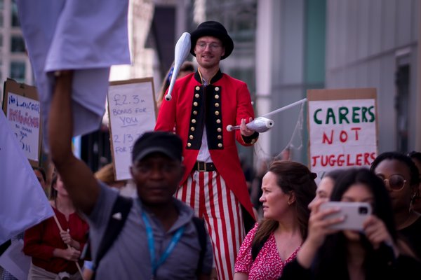 A juggler next to someone holding a sign saying 'care workers not jugglers' to call for a real Living Wage, Citizens UK action outside Barchester and Bupa, July 2022