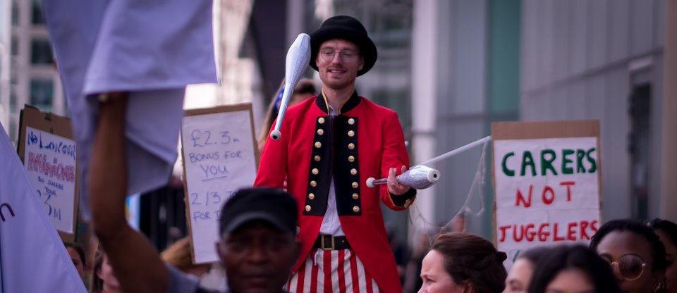 A juggler next to someone holding a sign saying 'care workers not jugglers' to call for a real Living Wage, Citizens UK action outside Barchester and Bupa, July 2022