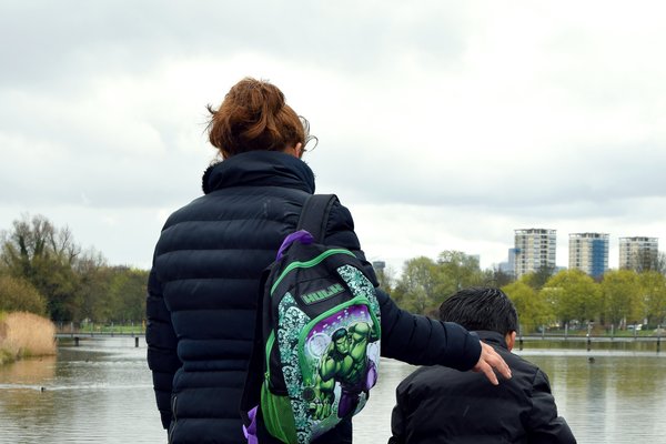 Back of a mother with her arm over her son, carrying his hulk backpack looking at a pond