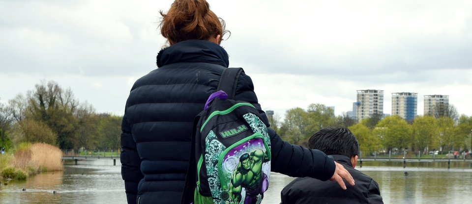 Back of a mother with her arm over her son, carrying his hulk backpack looking at a pond