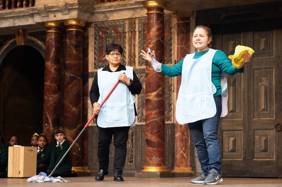 Two leaders stand on stage at Shakespeare's Globe, wearing aprons and holding cloths and a mop.