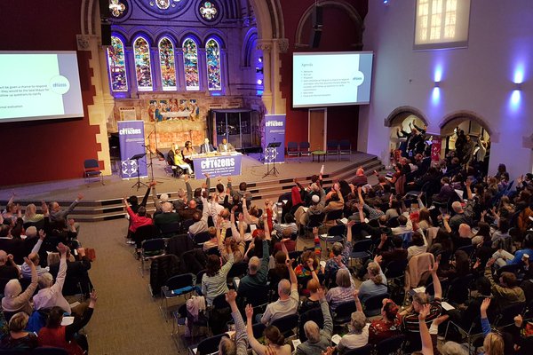 A large group of members are seated in a Church, as part of the Leicester and Leicestershire Citizens Founding Assembly. Their hands are all raised in solidarity