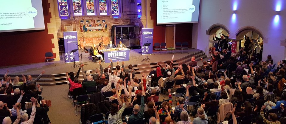 A large group of members are seated in a Church, as part of the Leicester and Leicestershire Citizens Founding Assembly. Their hands are all raised in solidarity