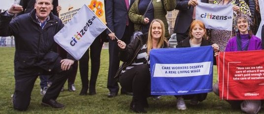 Citizens UK staff and leaders are gathered outside Parliament Square in Westminster, holding up flags which say "Care workers deserve a real Living Wage".