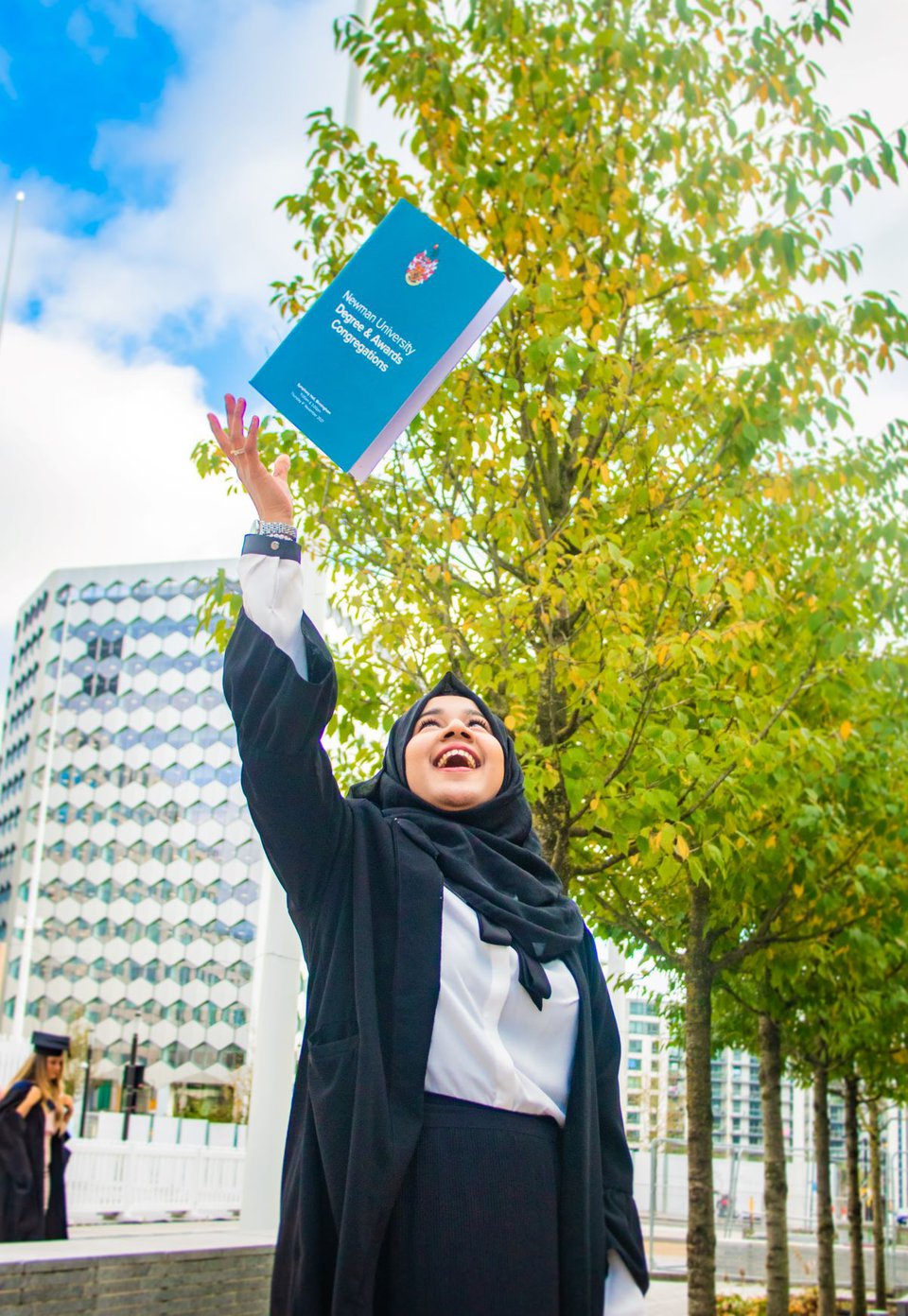 Woman stands in front of a row of trees and smiles throwing degree certification in the air