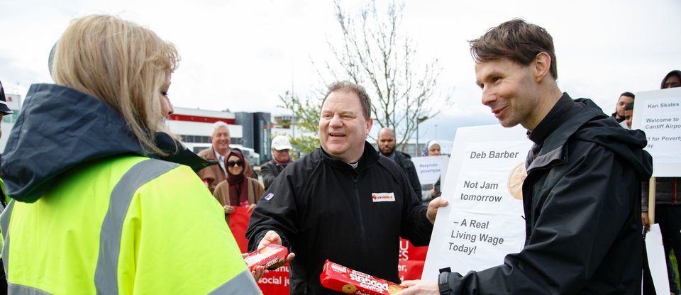 Priests hand jammy dodger biscuit packets to worker at Cardiff Airport at the Living Wage public action, Cymru Wales Citizens