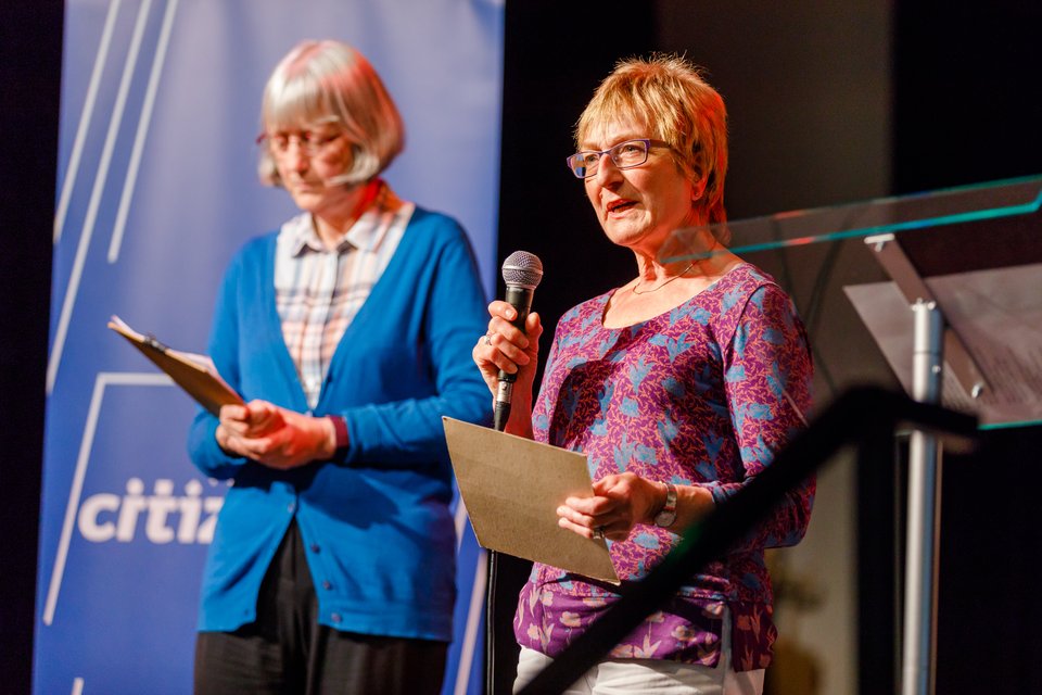 Two women stand on stage at a Citizens UK event, holding a microphone and speaking to the crowd