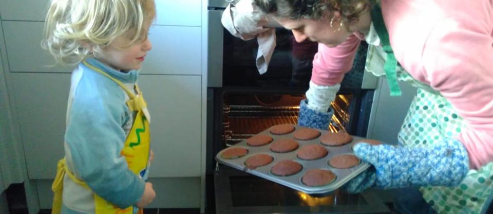 A young child looks on eagerly as a parent pulls out a tray of muffins that have been baking in the oven.