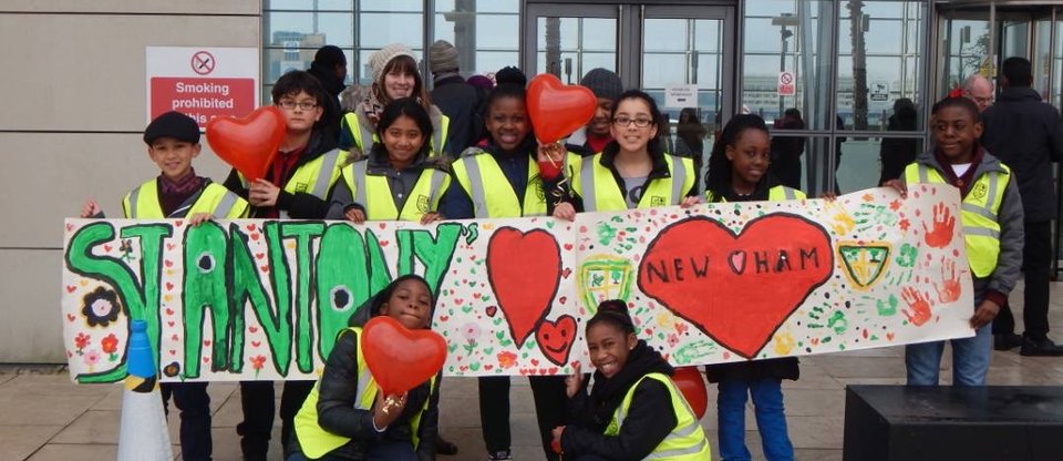 Children with high vis vests stand outside a building smiling and holding red love hearts. They also hold a hand drawn banner which says 'St Antony' to represent their school.