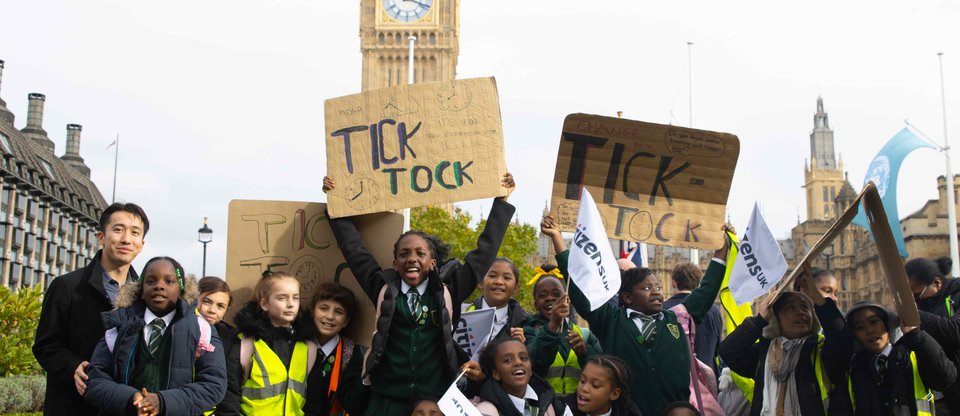 Young leaders in school uniforms stand infront of Big Ben at Parliament Square with 'Tik Tok' signs and Citizens UK flags, calling for better provisions around temporary accommodation