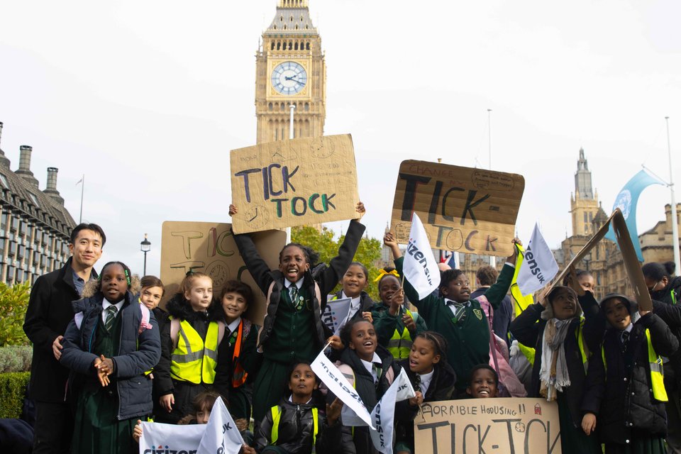 Young leaders in school uniforms stand infront of Big Ben at Parliament Square with 'Tik Tok' signs and Citizens UK flags, calling for better provisions around temporary accommodation