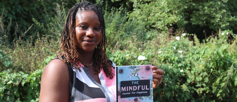 A woman (Saran) is smiling and holding a notebook that reads 'The mindful journal of happiness'