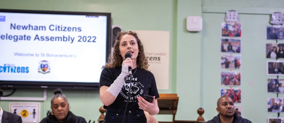 Young female leader speaking into a microphone at the Newham Citizens Delegates Assembly 2022
