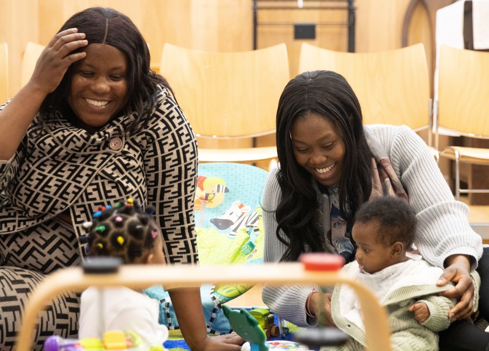 Two mums are smiling and playing with their toddlers.