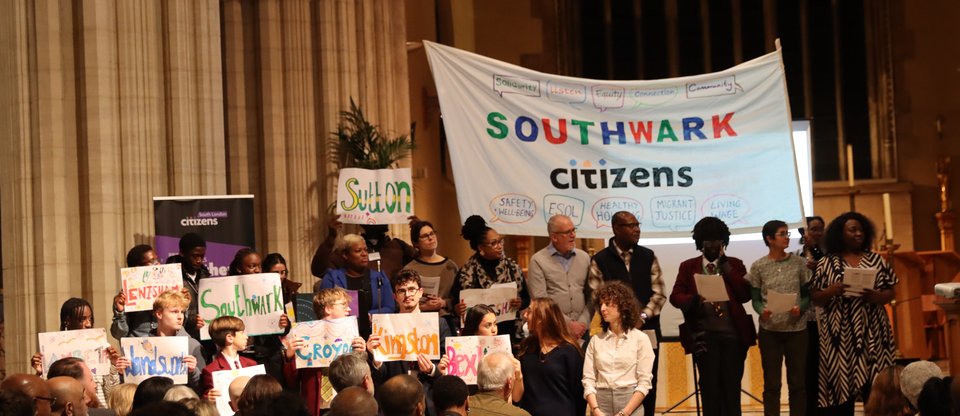 Many community leaders are on stage, a big banner with Southwark Citizens on it is behind them, and different community leaders are holding local signs that read Merton, Lewisham, etc