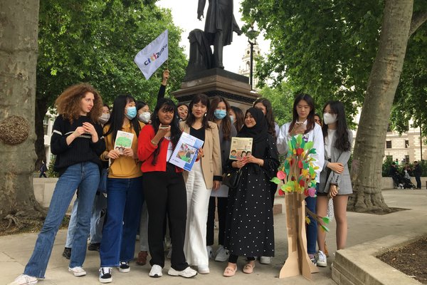 A group of young women stand outside waving a Citizens UK flag. They are celebrating a win for migration justice and have leaflets and a flowering tree sculpture.