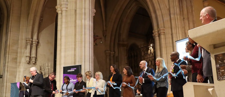 Local decision makers and community leaders stand on stage at the South London Citizens assembly with a long piece of connected strings