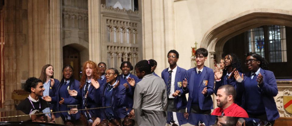 Children in school uniforms are standing behind a piano being played singing