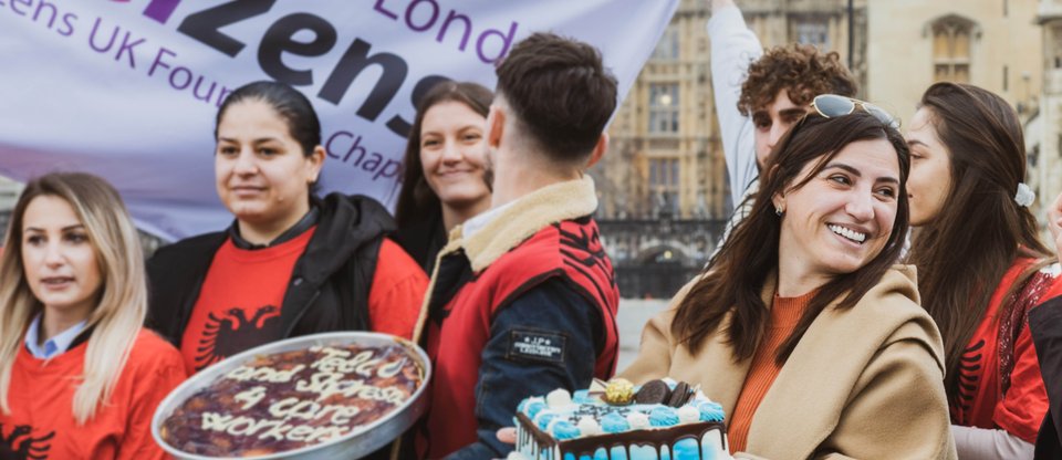 People standing with cakes and a Citizens UK flag. They are smiling and wearing shirts with the Albanian flag on it.