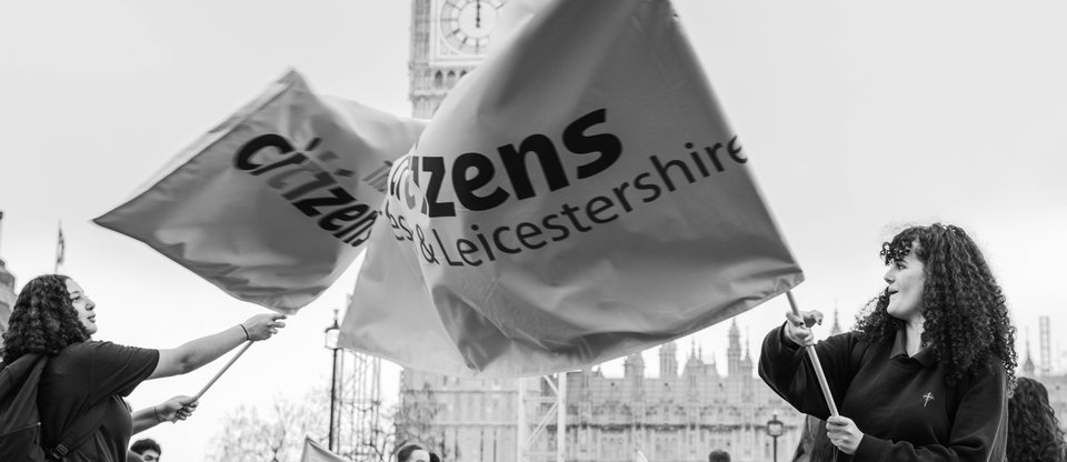 Black and white shot of two students waiving big Citizens UK flags on parliament square