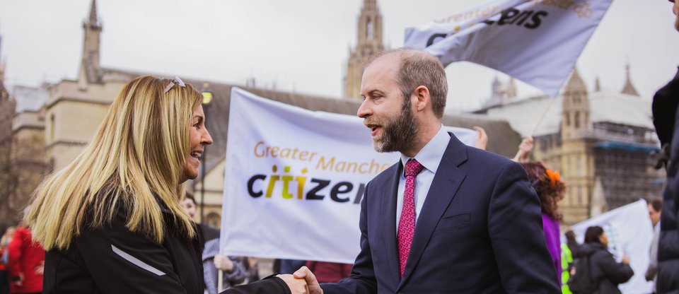 A Citizens UK leader smiles and shakes hands with an MP. In the background, a large Greater Manchester Citizens flag is flying.
