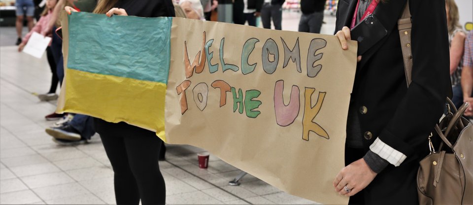 Две женщины стоят в зале прибытия в аэропорту с плакатом с украинским флагом, на котором написано: «Добро пожаловать в Великобританию»