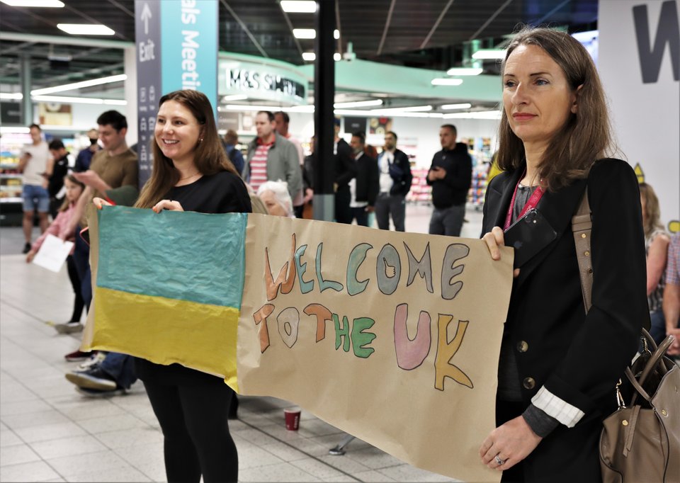 Two women stand at airport arrivals holding poster with Ukrainian flag saying "Welcome to the UK"