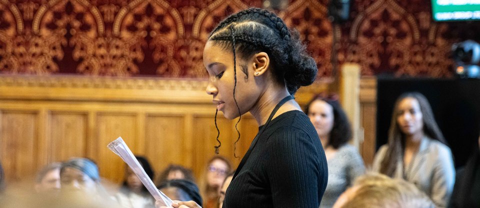 a young leader is speaking to a full room of attendees in Parliament holding a paper