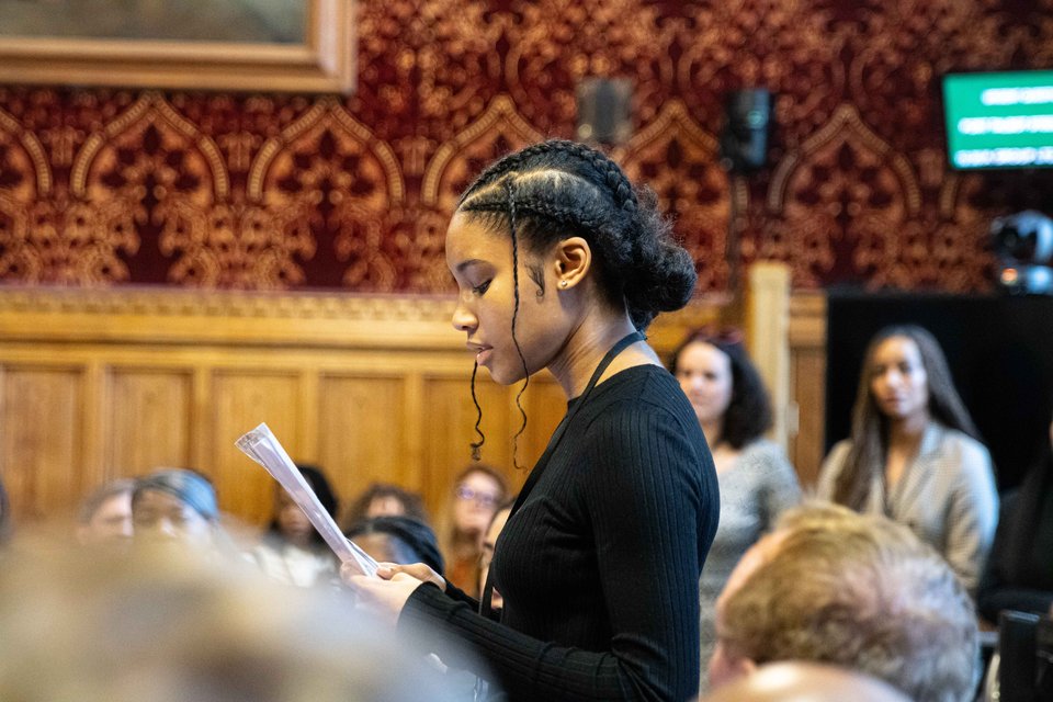 a young leader is speaking to a full room of attendees in Parliament holding a paper