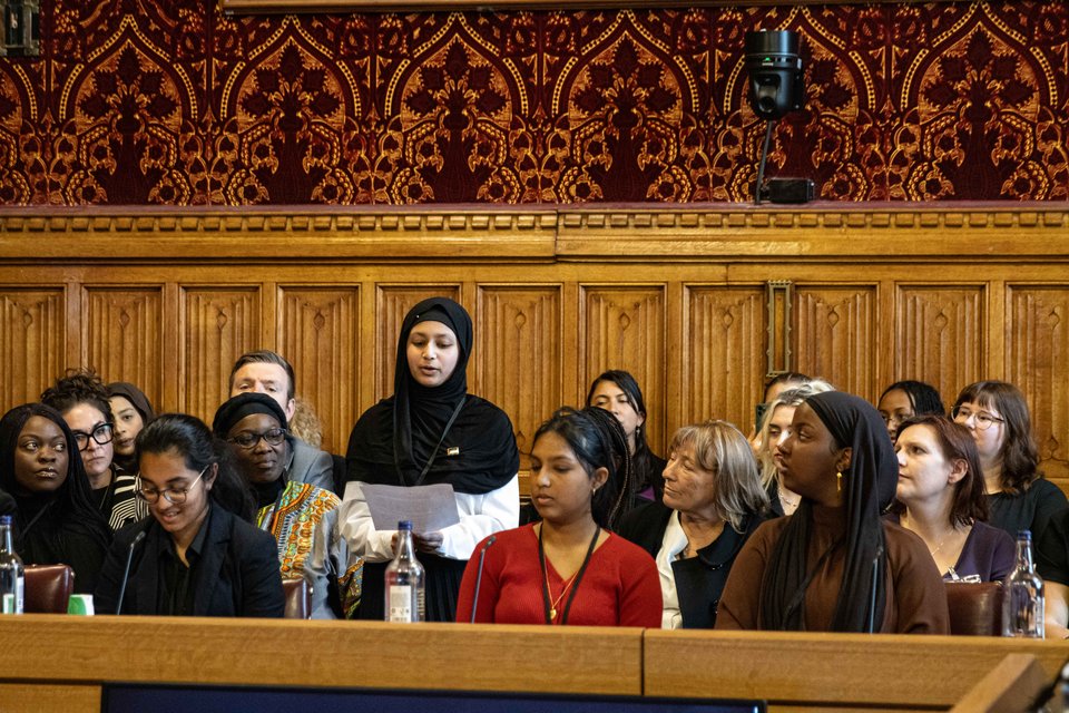 a girl in a hijab is standing speaking to a a room of seated attendees