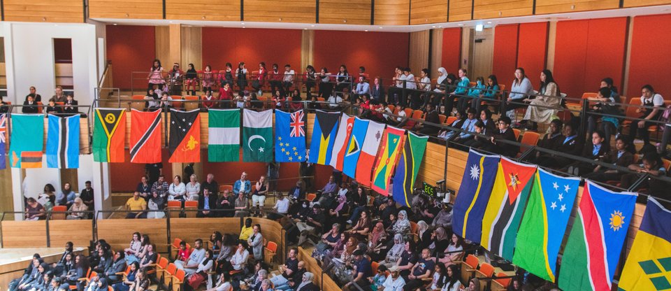A hall filled of people that make up Birmingham Citizens UK alliance. Around the hall, different flags of the Commonwealth nations hang together.