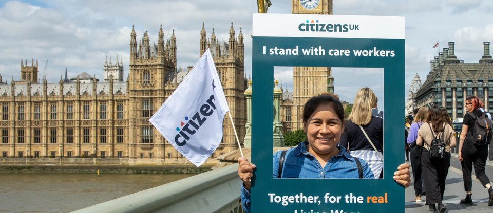 Person stands on Westminster Bridge with Parliament in the background, waving a Citizens UK flag and a selfie frame with support for the real Living Wage for carers