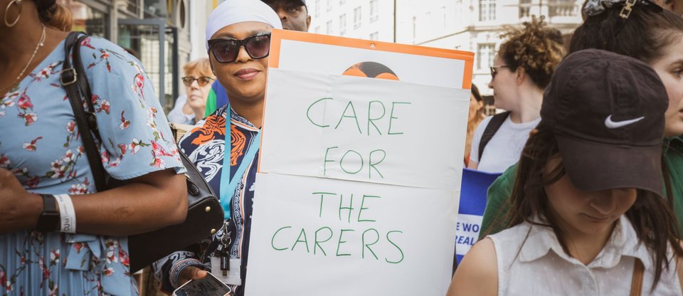 Person stands in the middle of a march holding a sign that says 'care for the carers'.