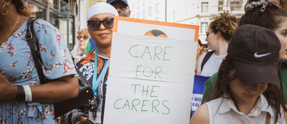 Person stands in the middle of a march holding a sign that says 'care for the carers'.