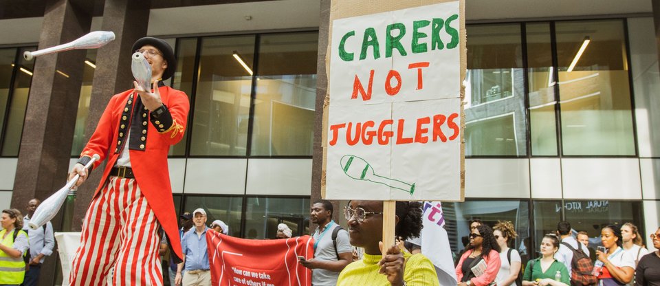 Juggler on stilts standing in front of people with sign "Carers not Jugglers" during Living Wage for Social Care public action on major care providers, Barchester and Bupa. July 2022