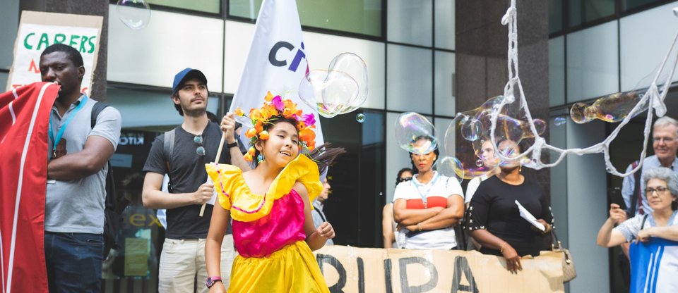 Young girl in yellow dress and with a flower headband dances. She's surrounded by giant bubbles and other community leaders holding signs and flags. One banner says 'Bupa do you care'. Image taken outside Bupa headquarters
